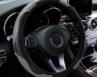 May include: A steering wheel cover featuring a combination of smooth gray panels and textured black carbon fiber-style sections. The cover is fitted onto a black steering wheel with integrated control buttons, positioned inside a vehicle interior.