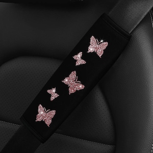 Puede incluir: Una funda para cinturón de seguridad negra con cinco aplicaciones de mariposas de color oro rosa brillante dispuestas en diagonal. Las mariposas varían en tamaño y están colocadas a lo largo de la funda de tela rectangular sobre el cinturón.