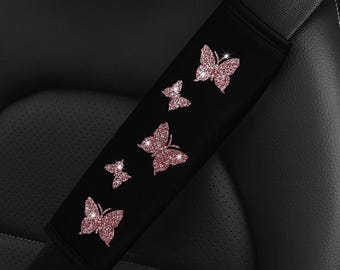 May include: A black seat belt cover featuring five glittery rose gold butterfly appliques arranged in a diagonal pattern. The butterflies vary in size and are positioned along the length of the rectangular fabric sleeve, which is secured over a seat belt.