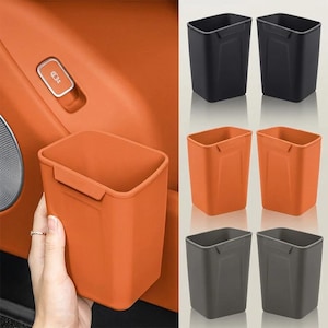 Puede incluir: Un contenedor de almacenamiento rectangular de parte superior abierta con un acabado liso y una pequeña pestaña en forma de gancho en el borde superior. El contenedor se muestra en negro, naranja y gris oscuro, con la versión naranja sostenida.