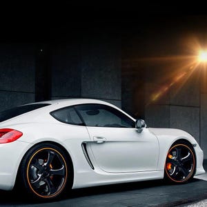 Puede incluir: Un coche deportivo blanco con una carrocer&iacute;a aerodin&aacute;mica, que presenta llantas negras de cinco radios con detalles en naranja en el borde y pinzas de freno de color naranja brillante. El veh&iacute;culo se muestra de perfil con un destello de luz arriba.