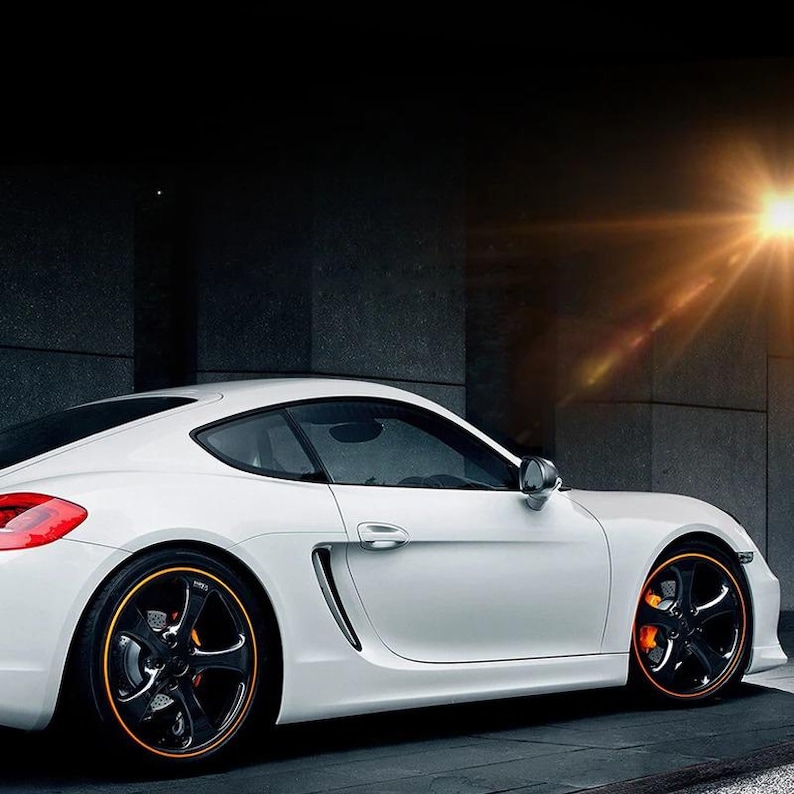 Puede incluir: Un coche deportivo blanco con una carrocer&iacute;a aerodin&aacute;mica, que presenta llantas negras de cinco radios con detalles en naranja en el borde y pinzas de freno de color naranja brillante. El veh&iacute;culo se muestra de perfil con un destello de luz arriba.