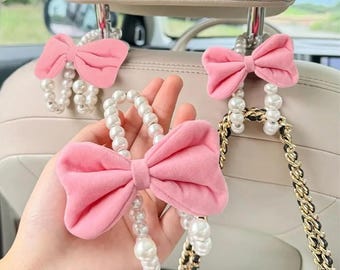 May include: A set of car headrest hooks featuring a pink fabric bow and a string of white faux pearls. The hooks are designed to hang from the metal posts of a vehicle headrest, providing a decorative storage solution for bags or accessories.
