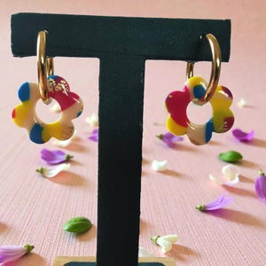May include: A pair of gold-toned hoop earrings featuring dangling flower-shaped charms. The charms are crafted from multi-colored segments in shades of pink, yellow, blue, and white, accented with small flecks of gold foil throughout the surface.
