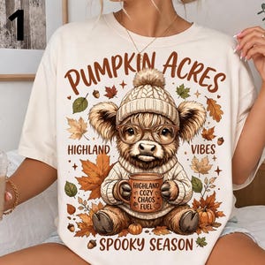Può includere: Una t-shirt color crema con la grafica di una mucca Highland che indossa un berretto di lana e occhiali, mentre tiene una tazza con l'etichetta 'HIGHLAND COZY CHAOS FUEL'. Il testo 'PUMPKIN ACRES', 'HIGHLAND VIBES' e 'SPOOKY SEASON' circonda la mucca.
