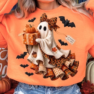 May include: An orange sweatshirt featuring a graphic of a ghost wearing a leopard print bow, holding a gift box with a pumpkin on top. The ghost is surrounded by black bats, autumn leaves, and an acorn, with a tag reading A PREPPY FALL GIFT.