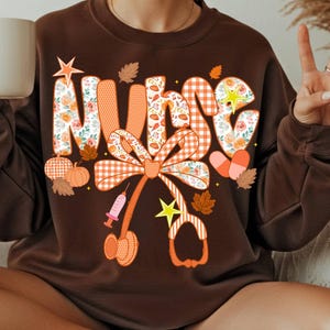 May include: A dark brown crewneck sweatshirt featuring the word NURSE in large, patterned letters. The design includes orange and white gingham, floral prints, pumpkins, autumn leaves, stars, a syringe, a stethoscope, and two pill capsules.