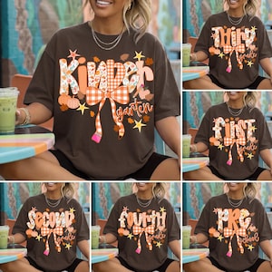 Puede incluir: Una camiseta de manga corta de color marrón oscuro con un estampado gráfico que incluye el texto Kindergarten, Third Grade, First Grade, Second Grade, Fourth Grade o Pre-K. El diseño presenta lazos de cuadros naranjas, hojas de otoño, calabazas, estrellas y lápices.