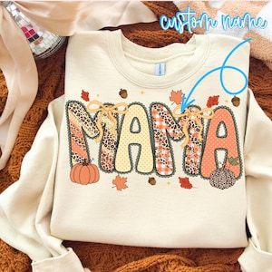 Può includere: Una felpa girocollo color crema con la parola MAMA in grandi lettere decorate. Le lettere presentano motivi leopardati, scacchi arancioni e pois, arricchite da fiocchi gialli, zucche, ghiande e foglie autunnali.