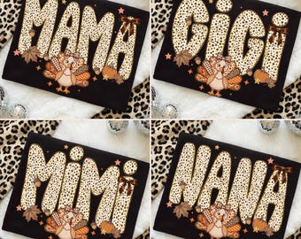 May include: A black t-shirt featuring a large graphic with the word MAMA, GIGI, MIMI, or NANA in a tan and brown leopard print. The design includes a cartoon turkey with a plaid bow, orange pumpkins, brown acorns, and small stars.