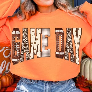 May include: A bright orange long-sleeve sweatshirt featuring the text "GAME DAY" across the chest. The letters are filled with a variety of patterns including a brown football texture, a black and tan leopard print, and a tan and white gingham check.