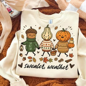 Könnte beinhalten: Ein cremefarbenes Sweatshirt mit Rundhalsausschnitt und einer Grafik von einer Eichel, einem Kürbis und einem Flaschenkürbis, die gemusterte Pullover tragen. Die Figuren halten eine Laterne und einen Korb. Der Text "sweater weather" steht darunter.