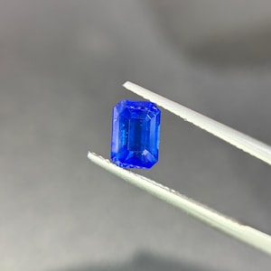 May include: A vibrant, deep blue gemstone with an emerald-cut shape, featuring a rectangular outline and faceted surfaces that reflect light. The stone is held by a pair of silver-toned metal tweezers, showcasing its clear, saturated colour and precise geometric cut.