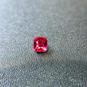 Puede incluir: Una piedra preciosa de talla cojín con un color rojo intenso y vibrante, con múltiples facetas que reflejan la luz. La piedra muestra una calidad clara y transparente con bordes definidos y una forma cuadrada simétrica y pulida.