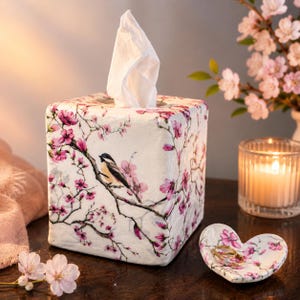 May include: A square tissue box cover and a heart-shaped trinket dish, both featuring a white base with a pattern of pink cherry blossoms and a small bird perched on a branch. A white tissue extends from the top of the box, and a gold ring sits inside the dish.