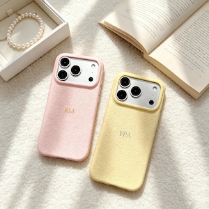 Op de afbeelding: Twee getextureerde telefoonhoesjes met camera-uitsparingen, één in lichtroze met het gouden monogram RM en één in lichtgeel met het zilveren monogram FPA. De hoesjes hebben een generfde afwerking en liggen naast een armband van parels.