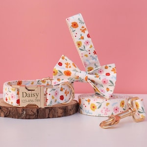 Puede incluir: Un conjunto de collar y correa para mascotas en color blanco con un estampado floral de flores naranjas, rojas y rosas. El collar incluye una hebilla de metal dorado grabada con el nombre Daisy y el número 123-456-789, además de un lazo a juego.