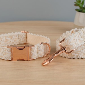 May include: A cream-coloured lace dog collar and matching lead set featuring rose gold metal hardware. The collar includes a side-release buckle and a D-ring, while the lead is equipped with a rose gold swivel snap hook for secure attachment.
