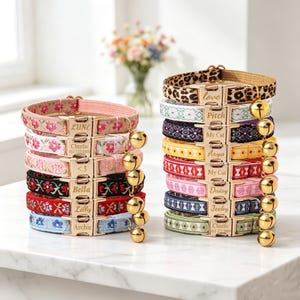 May include: Two stacks of pet collars featuring various woven patterns, including floral, geometric, and leopard print designs. Each collar has a gold-toned metal buckle engraved with names or text and a small gold-toned bell attached to the side.