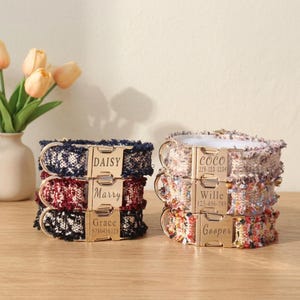 May include: Two stacks of pet collars featuring textured fabric in various colors including navy, burgundy, black, and multi-colored tweed. Each collar has a gold-toned metal buckle engraved with names like Daisy, Marry, Grace, Coco, Wille, and Gooper.