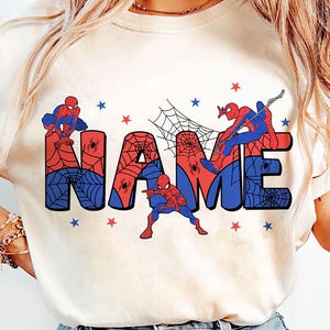 May include: A cream-colored short-sleeve t-shirt featuring a graphic print of the word NAME in large block letters. The letters are filled with a red and blue spiderweb pattern, accented by illustrations of a superhero and small red and blue stars.