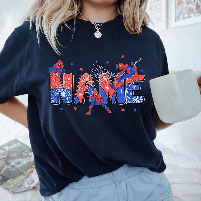 May include: A dark navy blue short-sleeve t-shirt featuring a graphic print of the word NAME in large block letters. The letters are filled with red and blue spiderweb patterns and accented with illustrations of a superhero in a red and blue suit.