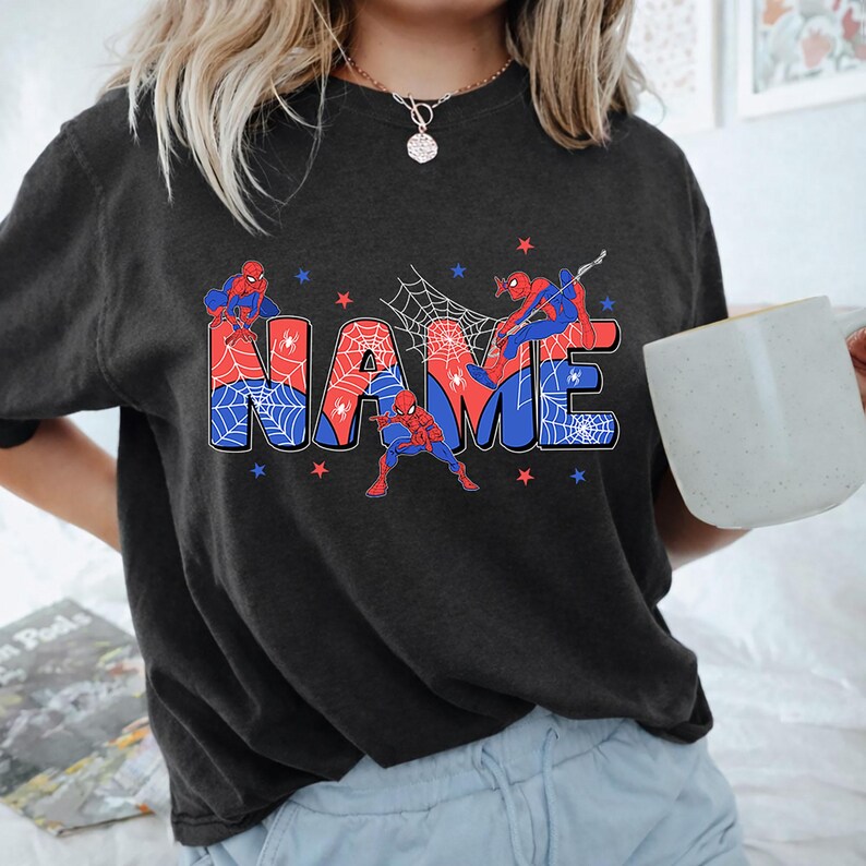 May include: A charcoal gray t-shirt featuring the word NAME in large, blocky letters filled with a red and blue spiderweb pattern. Three Spider-Man figures in red and blue suits are positioned around the text, accented by small red and blue stars.
