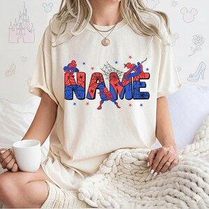 May include: A cream-colored short-sleeve t-shirt featuring a graphic print of the word NAME in large block letters. The letters are filled with a red and blue spiderweb pattern, accented by illustrations of a superhero character and small stars.