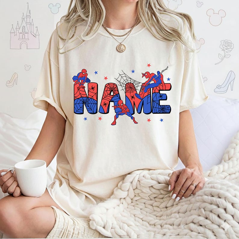May include: A cream-colored short-sleeve t-shirt featuring a graphic print of the word NAME in large block letters. The letters are filled with a red and blue spiderweb pattern, accented by illustrations of a superhero character and small stars.