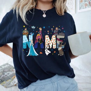 May include: A dark navy blue t-shirt featuring a graphic print of the word NAME in large block letters. The letters are decorated with characters and patterns from the movie Frozen, including Elsa, Anna, Olaf, Kristoff, and Sven, with snowflakes.
