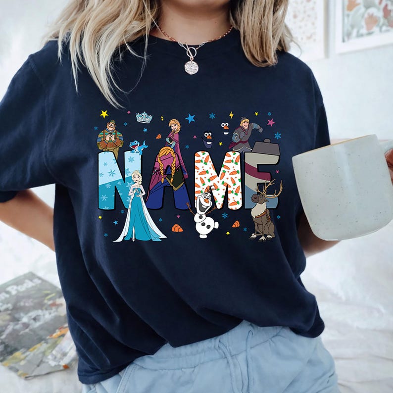 May include: A dark navy blue t-shirt featuring a graphic print of the word NAME in large block letters. The letters are decorated with characters and patterns from the movie Frozen, including Elsa, Anna, Olaf, Kristoff, and Sven, with snowflakes.
