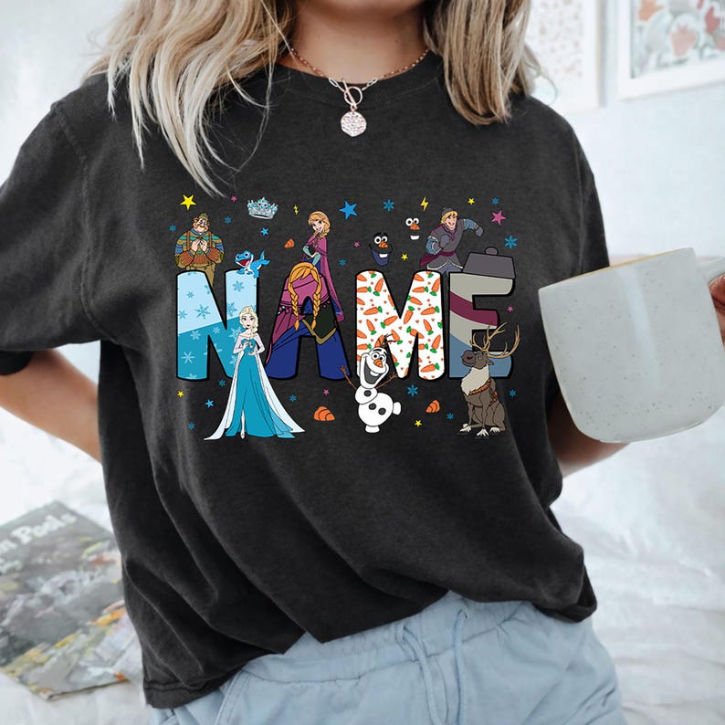 May include: A charcoal gray t-shirt featuring the word NAME in large block letters. The letters are decorated with Frozen characters including Elsa, Anna, Olaf, Kristoff, Sven, and Oaken, along with snowflake and carrot patterns and small stars.