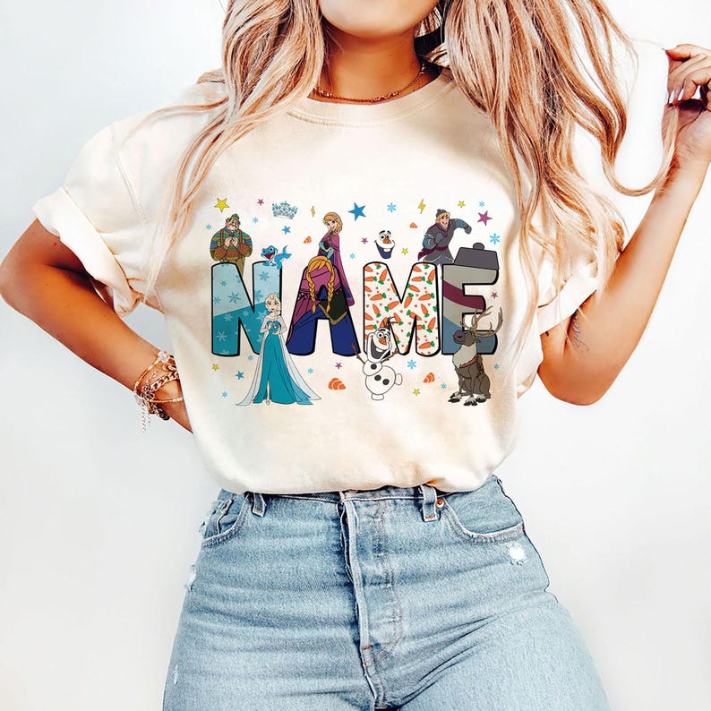 May include: A cream-colored short-sleeve t-shirt featuring the word NAME in large block letters. The letters are decorated with various characters from the movie Frozen, including Elsa, Anna, Olaf, Kristoff, and Sven, alongside snowflake and carrot patterns.