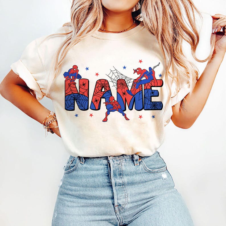 May include: A cream-colored short-sleeve t-shirt featuring a graphic print of the word NAME in large block letters. The letters are filled with a red and blue spiderweb pattern, accented by illustrations of a superhero and small red and blue stars.