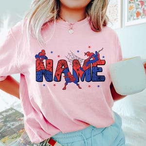 May include: A light pink t-shirt featuring a graphic design with the word NAME in large block letters filled with a red and blue spiderweb pattern. Three Spider-Man figures are positioned around the letters, accented by small red and blue stars.