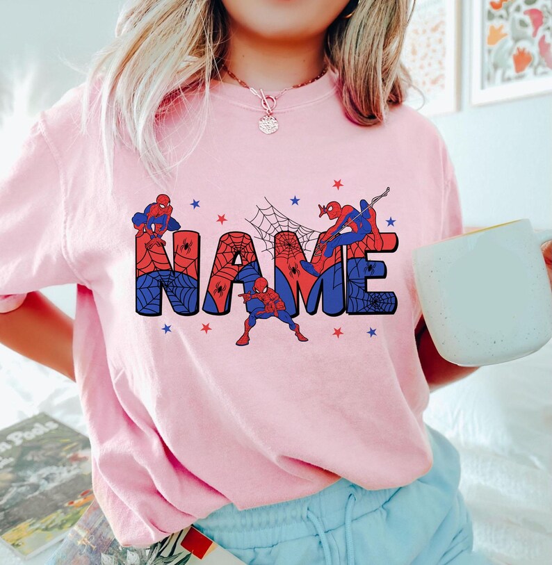 May include: A light pink t-shirt featuring a graphic design with the word NAME in large block letters filled with a red and blue spiderweb pattern. Three Spider-Man figures are positioned around the letters, accented by small red and blue stars.