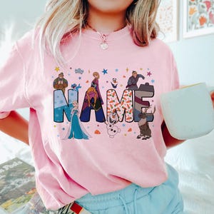 May include: A light pink t-shirt featuring the word NAME in large, colorful letters decorated with Frozen characters and motifs. The design includes Elsa, Anna, Olaf, Kristoff, and Sven, surrounded by small stars, snowflakes, and carrot icons.