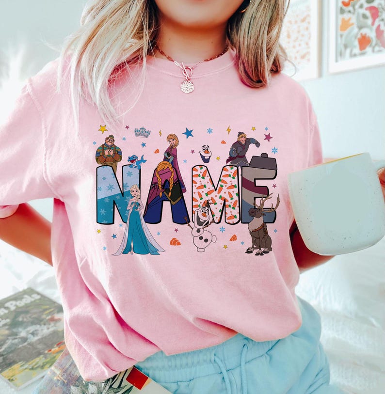 May include: A light pink t-shirt featuring the word NAME in large, colorful letters decorated with Frozen characters and motifs. The design includes Elsa, Anna, Olaf, Kristoff, and Sven, surrounded by small stars, snowflakes, and carrot icons.
