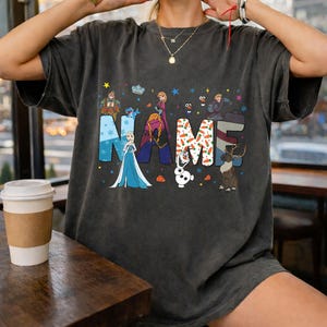 May include: A charcoal gray t-shirt featuring the word NAME in large block letters, each filled with different patterns and colors. The design includes illustrations of characters from the movie Frozen, including Elsa, Anna, Olaf, and Sven, surrounded by stars.