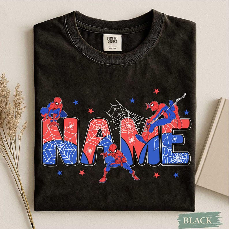 May include: A black crew neck t-shirt featuring the word NAME in large, bold letters filled with a red and blue spiderweb pattern. Three Spider-Man figures in red and blue suits are positioned around the text, accented by small red and blue stars.