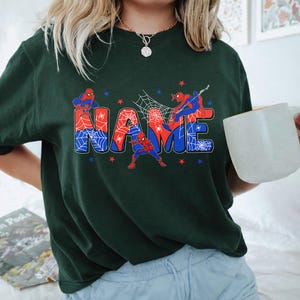 May include: A dark green t-shirt featuring a graphic print of the word NAME in large, blocky letters filled with a red and blue spiderweb pattern. Three red and blue superhero figures are positioned around the text, accented by small stars.