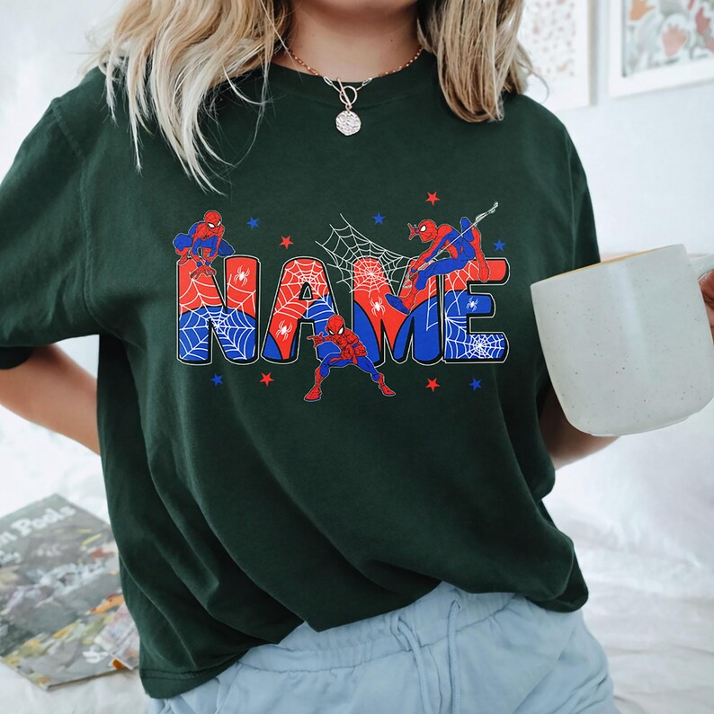 May include: A dark green t-shirt featuring a graphic print of the word NAME in large, blocky letters filled with a red and blue spiderweb pattern. Three red and blue superhero figures are positioned around the text, accented by small stars.