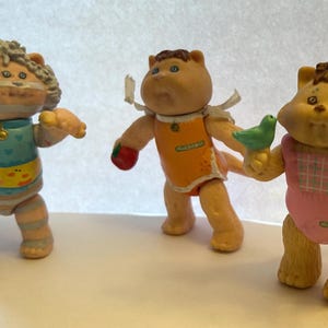 Op de afbeelding: Drie vintage Cabbage Patch Kids beeldjes. De figuren zijn lichtbruin met verschillende outfits. Eén draagt een blauw en geel outfit, een ander een oranje outfit en de derde een roze outfit. Eén figuur houdt een groene vogel vast en een ander een rode appel.