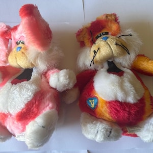 Puede incluir: Dos figuras de peluche de un gato de dibujos animados. Una es principalmente rosa y blanca, la otra es roja, naranja y amarilla. Ambas tienen pelaje blanco alrededor de la cara y el pecho, con ojos azules y bigotes negros. Los juguetes están tumbados.