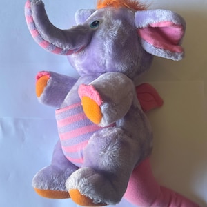 Op de afbeelding: Een paarse pluchen olifant met oranje en roze accenten. De olifant heeft een plukje oranje haar, roze gevoerde oren en een gestreepte buik. Zijn voeten en de binnenkant van zijn oren zijn oranje. De slurf is roze en wit gestreept.