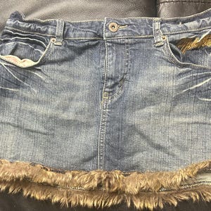 May include: A denim mini skirt with a frayed, faux fur trim at the hem. The skirt has classic five-pocket styling, a button closure, and belt loops. The denim is a medium wash with subtle fading.