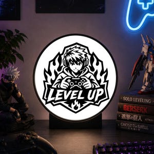 以下が含まれることがあります： ヘッドフォンを装着しゲームコントローラーを持つ人物の白黒グラフィックが描かれた円形のLEDライト。中央下部には「LEVEL UP」の文字があり、デザインは様式化された炎で縁取られ、黒い台座に取り付けられています。