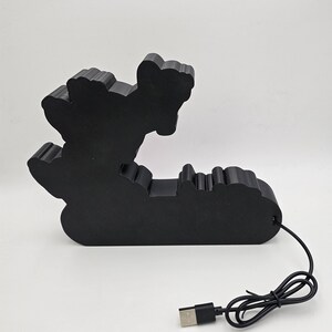 Puede incluir: Una l&aacute;mpara decorativa de color negro mate con una forma personalizada que presenta un dise&ntilde;o de silueta. El objeto tiene una superficie s&oacute;lida y opaca, e incluye un cable de alimentaci&oacute;n negro con un conector USB-A est&aacute;ndar en la base.