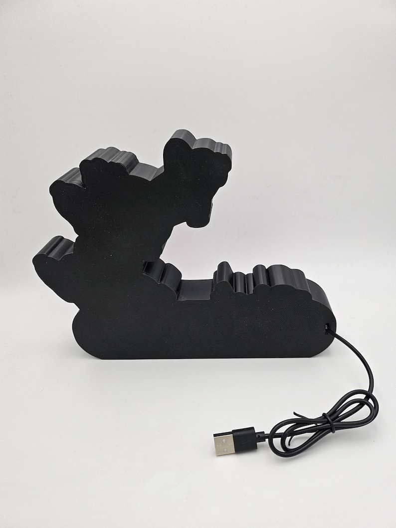 Puede incluir: Una l&aacute;mpara decorativa de color negro mate con una forma personalizada que presenta un dise&ntilde;o de silueta. El objeto tiene una superficie s&oacute;lida y opaca, e incluye un cable de alimentaci&oacute;n negro con un conector USB-A est&aacute;ndar en la base.