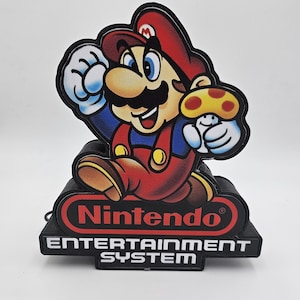 May include: A decorative light featuring Mario holding a yellow mushroom. The base is black with the red Nintendo logo and the white text ENTERTAINMENT SYSTEM. The character is depicted in a classic red and blue outfit with a brown hat and moustache.